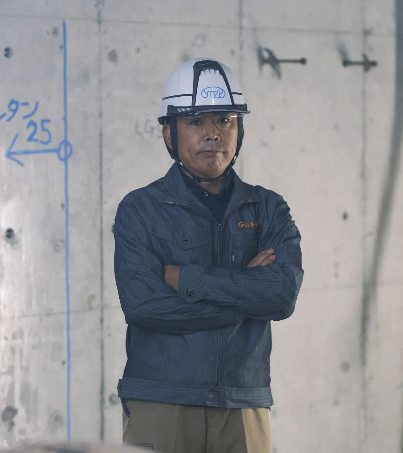 ヘルメットを着用し、腕を組んで真剣な表情で立つ作業着姿の男性